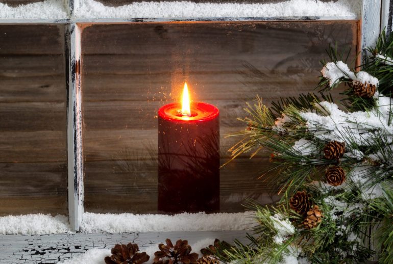 Decorated window with glowing red candle, selective focus on flame and top part of candle, pine tree, cones and snow outside. Christmas concept.
